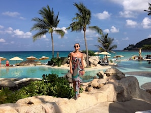 A model in a flowing white beach dress standing by a sun-drenched poolside with palm trees swaying gently.