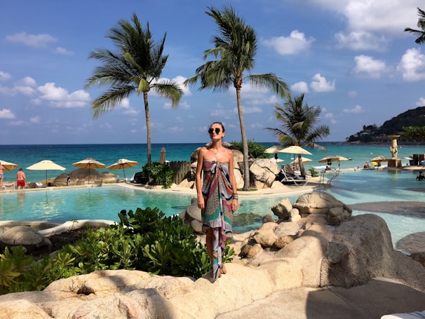 A woman wearing a vibrant tropical print dress standing on a sunny beach with palm trees in the background.