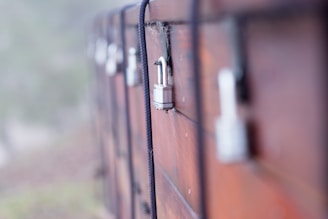 A series of weathered wooden lockers secured with metal padlocks and black ropes. The focus is on one padlock in the foreground, with a few blurred in the background. The surface of the wood appears rough with visible texture and some cobwebs, indicating age and outdoor exposure.