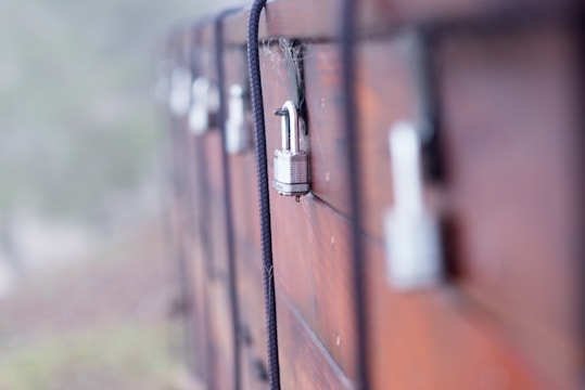A series of weathered wooden lockers secured with metal padlocks and black ropes. The focus is on one padlock in the foreground, with a few blurred in the background. The surface of the wood appears rough with visible texture and some cobwebs, indicating age and outdoor exposure.