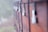 A series of weathered wooden lockers secured with metal padlocks and black ropes. The focus is on one padlock in the foreground, with a few blurred in the background. The surface of the wood appears rough with visible texture and some cobwebs, indicating age and outdoor exposure.