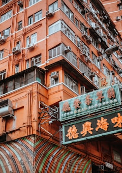 A vibrant, orange-colored building with multiple windows featuring air conditioning units on each. The structure includes traditional neon signs with Chinese characters, adding an authentic urban feel. The lower part shows a rounded, striped awning.