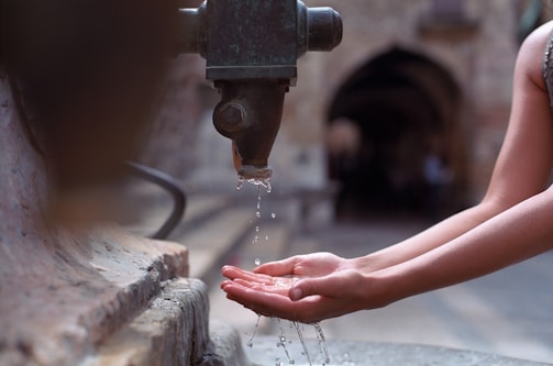 A pair of hands is positioned under an old metal faucet, catching drops of water. The background is slightly blurred, showing an outdoor setting with stone architecture.