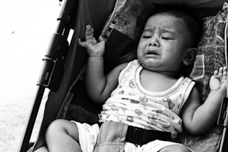 A baby sitting in a stroller appears to be crying. The expression on the baby's face shows distress, with tears visible and the mouth open as if wailing. The baby is wearing a sleeveless top and is secured with a harness in the stroller.