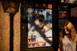 A young woman with long blonde hair wearing sunglasses and headphones walks past a magazine stand. The magazine cover prominently displays a glamorous model with an elegant dress, featuring various headlines related to fashion and city life in Paris. The lighting casts warm shadows on the scene, contributing to an overall sunlit ambiance.