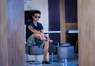 A person sitting casually on a modern stool in a contemporary indoor setting, wearing sunglasses, a black t-shirt, and shorts. The environment features wooden elements and neutral-toned walls, creating a relaxed atmosphere.