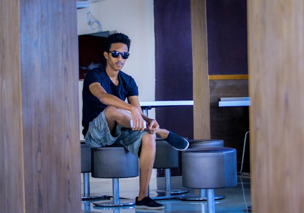 A person sitting casually on a modern stool in a contemporary indoor setting, wearing sunglasses, a black t-shirt, and shorts. The environment features wooden elements and neutral-toned walls, creating a relaxed atmosphere.