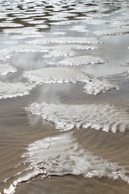 Wet sand patterns formed by waves, creating ripples and smooth textures. The surface glistens under light, reflecting a silvery tone with intricate natural designs.