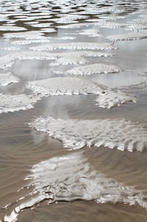 Wet sand patterns formed by waves, creating ripples and smooth textures. The surface glistens under light, reflecting a silvery tone with intricate natural designs.