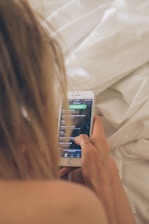 A person with long blonde hair is holding a smartphone displaying a music app interface, while sitting on a bed with white linens. The phone screen shows a playlist with song titles, and the person's hand is adorned with a ring.