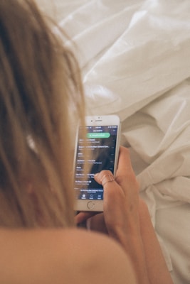A person with long blonde hair is holding a smartphone displaying a music app interface, while sitting on a bed with white linens. The phone screen shows a playlist with song titles, and the person's hand is adorned with a ring.