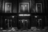 A black and white image featuring the front facade of a building with a neon sign reading 'Cafe Brauer'. The architecture includes brickwork and multiple doors with glass panels. Two illuminated bulbs are visible, adding a vintage ambiance.