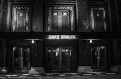 A black and white image featuring the front facade of a building with a neon sign reading 'Cafe Brauer'. The architecture includes brickwork and multiple doors with glass panels. Two illuminated bulbs are visible, adding a vintage ambiance.