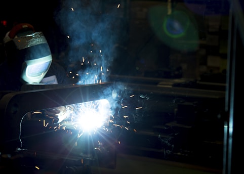 A welder wearing a protective mask is engaged in welding, producing bright sparks and a plume of smoke in a dimly lit industrial environment. The intense white light from the welding torch creates stark contrasts against the surrounding darkness.