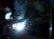 A welder wearing a protective mask is engaged in welding, producing bright sparks and a plume of smoke in a dimly lit industrial environment. The intense white light from the welding torch creates stark contrasts against the surrounding darkness.