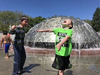 Children are joyfully playing in front of a large water fountain on a sunny day. One child is wearing a bright green shirt with 'Skate' written on it, and he has his arms outstretched, laughing as water splashes around him. Another child is nearby, similarly enjoying the playful environment. The background is filled with green trees under a clear blue sky.