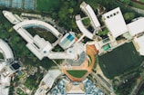 An aerial view of a large campus or complex featuring multiple modern buildings with various architectural designs, surrounded by greenery and athletic fields. The image shows a mixture of curved and angular structures along with roads and pathways connecting them. A football field with visible lines and several tennis courts are also present, providing recreational spaces within the area.