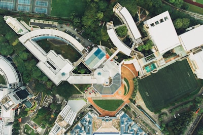 An aerial view of a large campus or complex featuring multiple modern buildings with various architectural designs, surrounded by greenery and athletic fields. The image shows a mixture of curved and angular structures along with roads and pathways connecting them. A football field with visible lines and several tennis courts are also present, providing recreational spaces within the area.