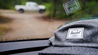 A gray hat with a Herschel Supply Co. brand label is placed on the dashboard of a vehicle, partially reflected in the windshield. The background shows a blurred view of an outdoor setting with a white pickup truck.