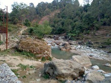 A serene, forested landscape with a shallow river running through rocky terrain. Large boulders and stones are scattered across the riverbank, surrounded by dense green vegetation and tall trees. A dirt path runs along the left side, partially hidden by foliage, with some power lines visible.