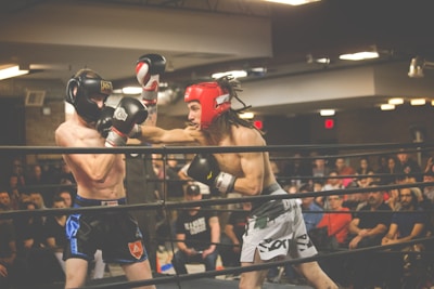 Two boxers are engaged in an intense match inside a boxing ring, one wearing a black headgear and the other in red. The audience is present in the background, watching the fight with interest. The scene is lively with bright overhead lights illuminating the setting.