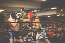 Two boxers are engaged in an intense match inside a boxing ring, one wearing a black headgear and the other in red. The audience is present in the background, watching the fight with interest. The scene is lively with bright overhead lights illuminating the setting.