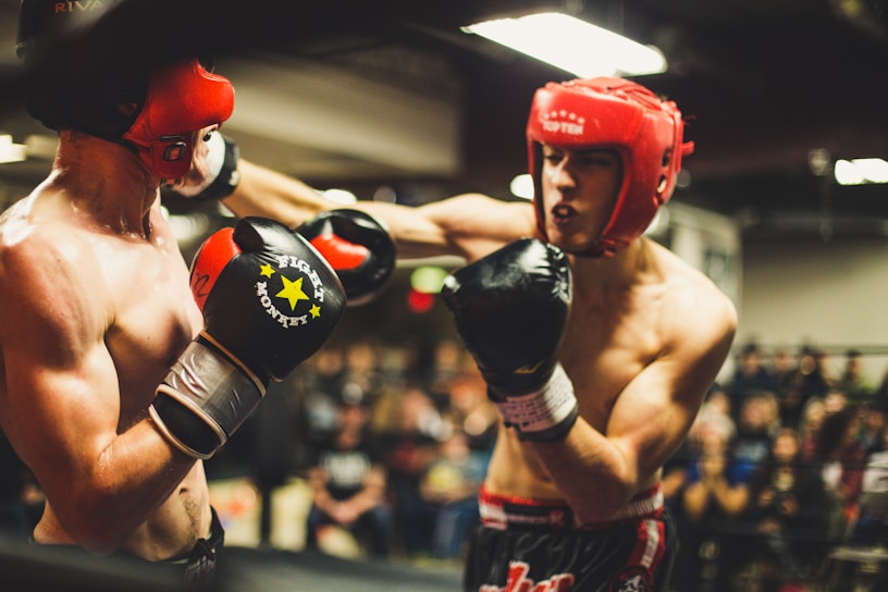 Two individuals are engaged in a boxing match. Both fighters are wearing red headgear and boxing gloves. One boxer is delivering a punch to the other's face. The background shows a blurred crowd of spectators watching the fight in a dimly lit arena.