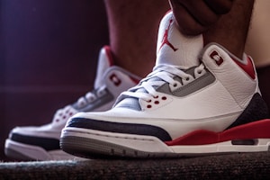 A close-up view of stylish sneakers with white leather, red accents, and a visible logo on the tongue. The shoes are being worn, with one hand adjusting the tongue.