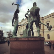 Three large bronze statues of muscular men holding hammers are prominently displayed against the backdrop of a classical building. The statues are positioned on a pedestal with inscriptions on its base. This setting is located in an urban environment with pedestrians and vehicles visible in the background.