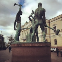 Three large bronze statues of muscular men holding hammers are prominently displayed against the backdrop of a classical building. The statues are positioned on a pedestal with inscriptions on its base. This setting is located in an urban environment with pedestrians and vehicles visible in the background.