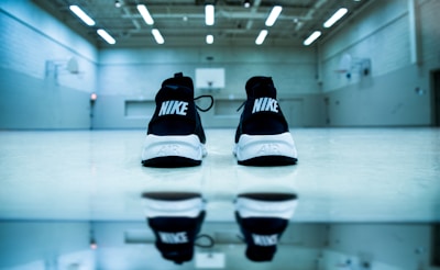 A pair of Nike sneakers is placed on a basketball court, with their backs facing the camera. The sneakers are black with white soles and are prominently featured against the smooth floor. The reflection of the sneakers is visible on the polished surface. The background shows a basketball court with hoops and high ceilings illuminated by fluorescent lights.