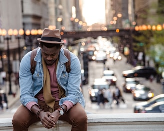 A man in a leather jacket looking down while sitting on a ledge in a city