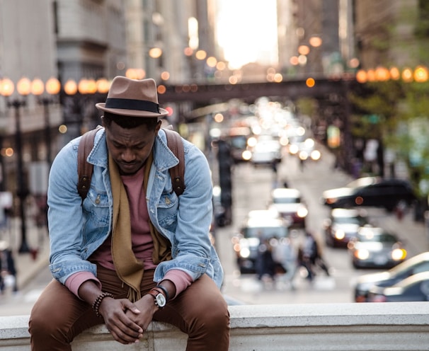 A man in a leather jacket looking down while sitting on a ledge in a city