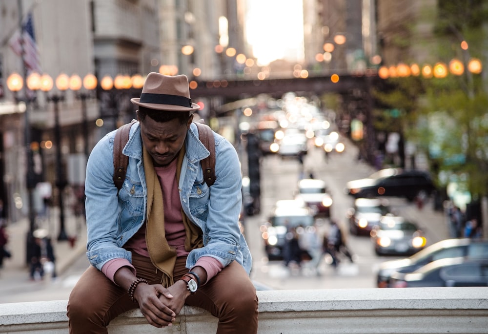 A man in a leather jacket looking down while sitting on a ledge in a city