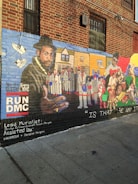 A detailed mural painted on the side of a brick building. The artwork features a large portrait of a man wearing a hat and gold chain on the left, with a background that includes various people in white suits and urban scenery. On the right side, another man is depicted alongside various colorful elements and figures. The mural includes text such as 'RUNDMC' and mentions the lead muralist and assistants. The lower part of the mural has the phrase 'IS THAT THE WAY IT IS.'