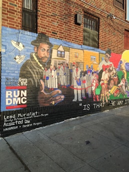 A detailed mural painted on the side of a brick building. The artwork features a large portrait of a man wearing a hat and gold chain on the left, with a background that includes various people in white suits and urban scenery. On the right side, another man is depicted alongside various colorful elements and figures. The mural includes text such as 'RUNDMC' and mentions the lead muralist and assistants. The lower part of the mural has the phrase 'IS THAT THE WAY IT IS.'