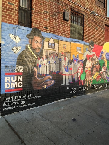 A detailed mural painted on the side of a brick building. The artwork features a large portrait of a man wearing a hat and gold chain on the left, with a background that includes various people in white suits and urban scenery. On the right side, another man is depicted alongside various colorful elements and figures. The mural includes text such as 'RUNDMC' and mentions the lead muralist and assistants. The lower part of the mural has the phrase 'IS THAT THE WAY IT IS.'