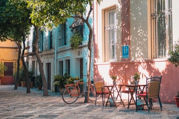 Sunlit street lined with colorful buildings and a cobblestone path. A quaint outdoor seating area features wooden tables and wicker chairs, surrounded by potted plants and bicycles. Shadows of leafy trees add a charming play of light.
