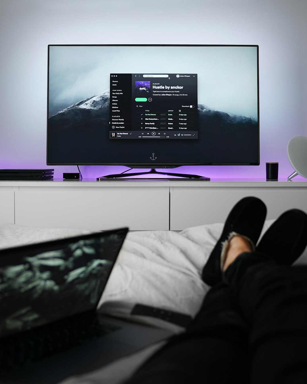 Person relaxing on bed with slippers listening to spotify through a television screen on a dresser