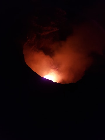 A deep crater with a bright, glowing orange and red light emanates from its center, suggesting volcanic activity or a large fire. The surrounding area is dark, emphasizing the illumination from within the crater.