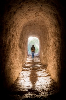 A person with a backpack walks through a rugged, earthy tunnel with light streaming in from an opening at the end. The rough texture of the tunnel walls suggests it might be a natural or ancient passageway, casting a long shadow on the floor. Lush greenery is visible outside the tunnel, illuminated by natural light.