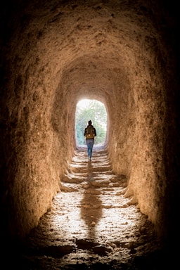 A person with a backpack walks through a rugged, earthy tunnel with light streaming in from an opening at the end. The rough texture of the tunnel walls suggests it might be a natural or ancient passageway, casting a long shadow on the floor. Lush greenery is visible outside the tunnel, illuminated by natural light.