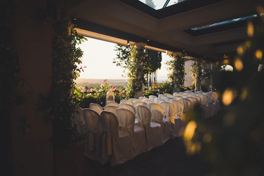 Rows of neatly arranged white chairs around wooden banquet tables, decorated with colorful table runners.