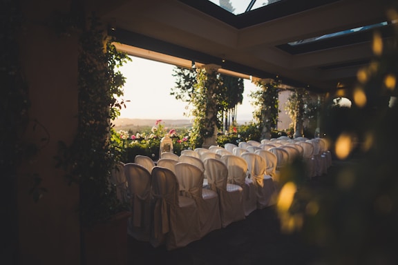 Rows of white chairs with seat covers are arranged facing a view of a scenic outdoor landscape. The setting is decorated with lush green foliage and floral arrangements. The space is illuminated with warm, natural light from large windows or an open ceiling.