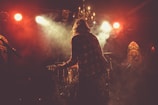 In a dimly lit venue, a musician with long hair and a checkered shirt stands on stage with a guitar, surrounded by thick smoke. Bright spotlights shine down, creating a dramatic atmosphere. A chandelier hangs from the ceiling, and stained glass panels decorate the background.