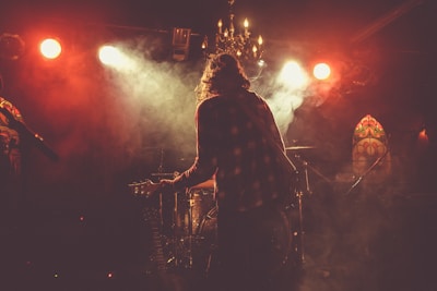In a dimly lit venue, a musician with long hair and a checkered shirt stands on stage with a guitar, surrounded by thick smoke. Bright spotlights shine down, creating a dramatic atmosphere. A chandelier hangs from the ceiling, and stained glass panels decorate the background.