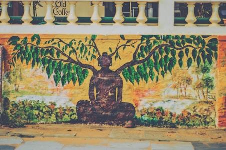 A mural on a wall features a serene silhouette of a person meditating beneath an abstract tree with lush green leaves. The background is a warm yellow with hints of orange and green depicting a nature-inspired scene. Above the artwork, a portion of a building is visible with the text 'Old Coffee'.