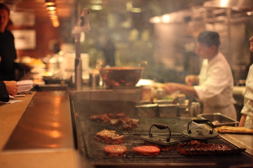 A busy kitchen setting with a chef preparing food on a grill. Various types of meat are being cooked, and a variety of kitchen implements and ingredients are visible. The ambiance is warm and dimly lit, adding to the bustling yet cozy atmosphere.