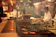 A busy kitchen setting with a chef preparing food on a grill. Various types of meat are being cooked, and a variety of kitchen implements and ingredients are visible. The ambiance is warm and dimly lit, adding to the bustling yet cozy atmosphere.