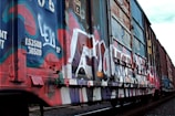 A freight train car covered in vibrant graffiti artwork is seen on a section of railway track. The graffiti features a mix of colors such as blue, red, and white, with various tags and designs painted over the entire side of the car. The surrounding environment appears industrial, and the train car shows signs of wear and tear.